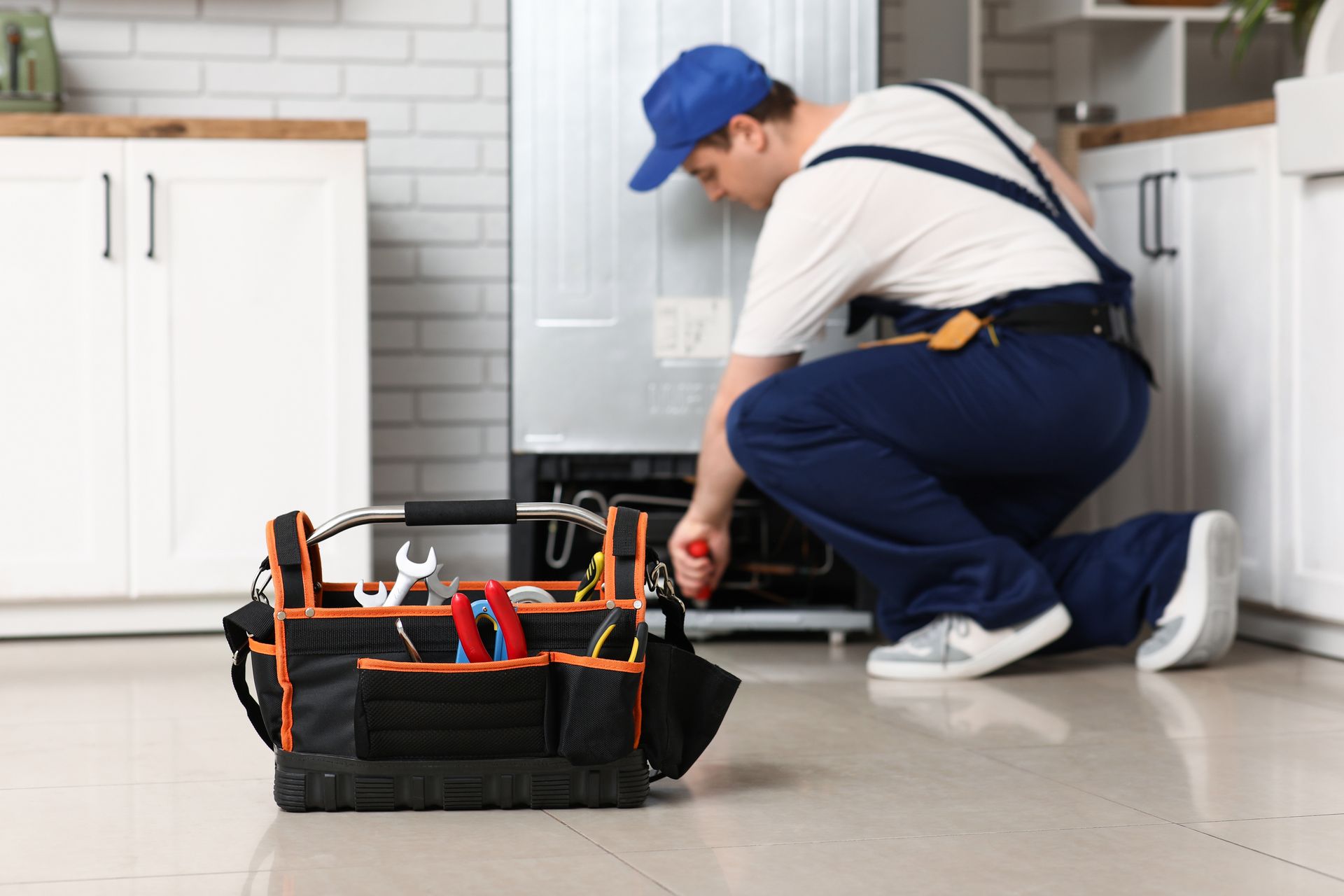 A technician in a blue cap and work uniform kneels on a kitchen floor, repairing the back of a refrigerator near a tool bag.