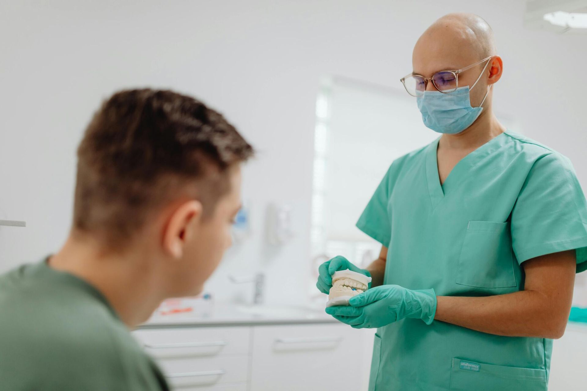 Dentist in green scrubs and blue mask preparing a syringe while speaking to a seated patient