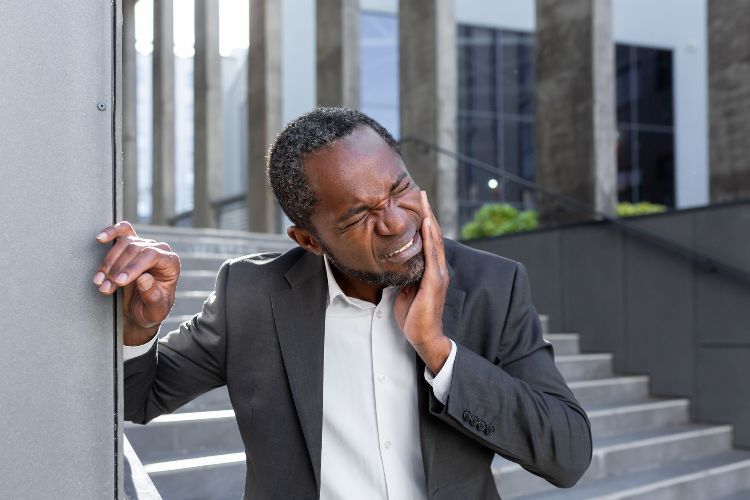 Businessman rubbing his cheek outdoors by office steps, looking uncomfortable while holding a phone.