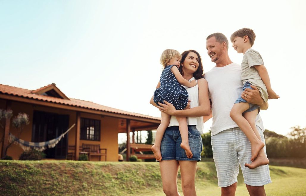 Family of four standing on grass in front of a rustic house, smiling and hugging in warm sunlight