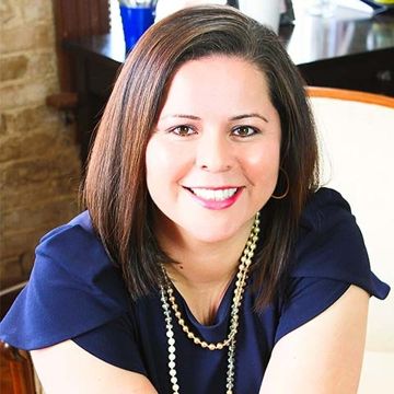 Smiling woman in a navy top and pearl necklace, seated indoors against a softly lit background