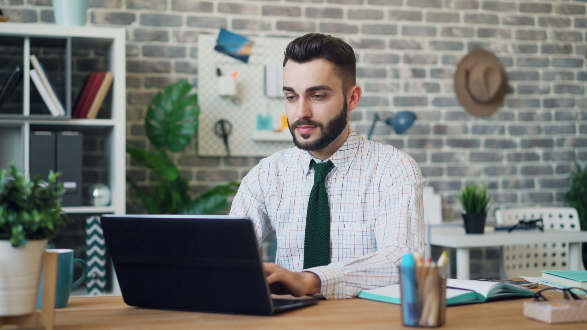 Man in white shirt and green tie working on a laptop at a desk in a brick-walled office