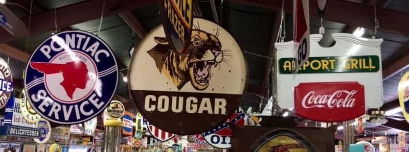 Vintage signs, including Pontiac Service, Cougar, and Coca-Cola, hang from the ceiling in an indoor collection.
