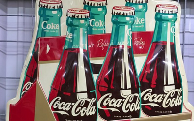 A cardboard display for Coca-Cola, featuring multiple illustrated glass bottles with vintage branding on a red background.
