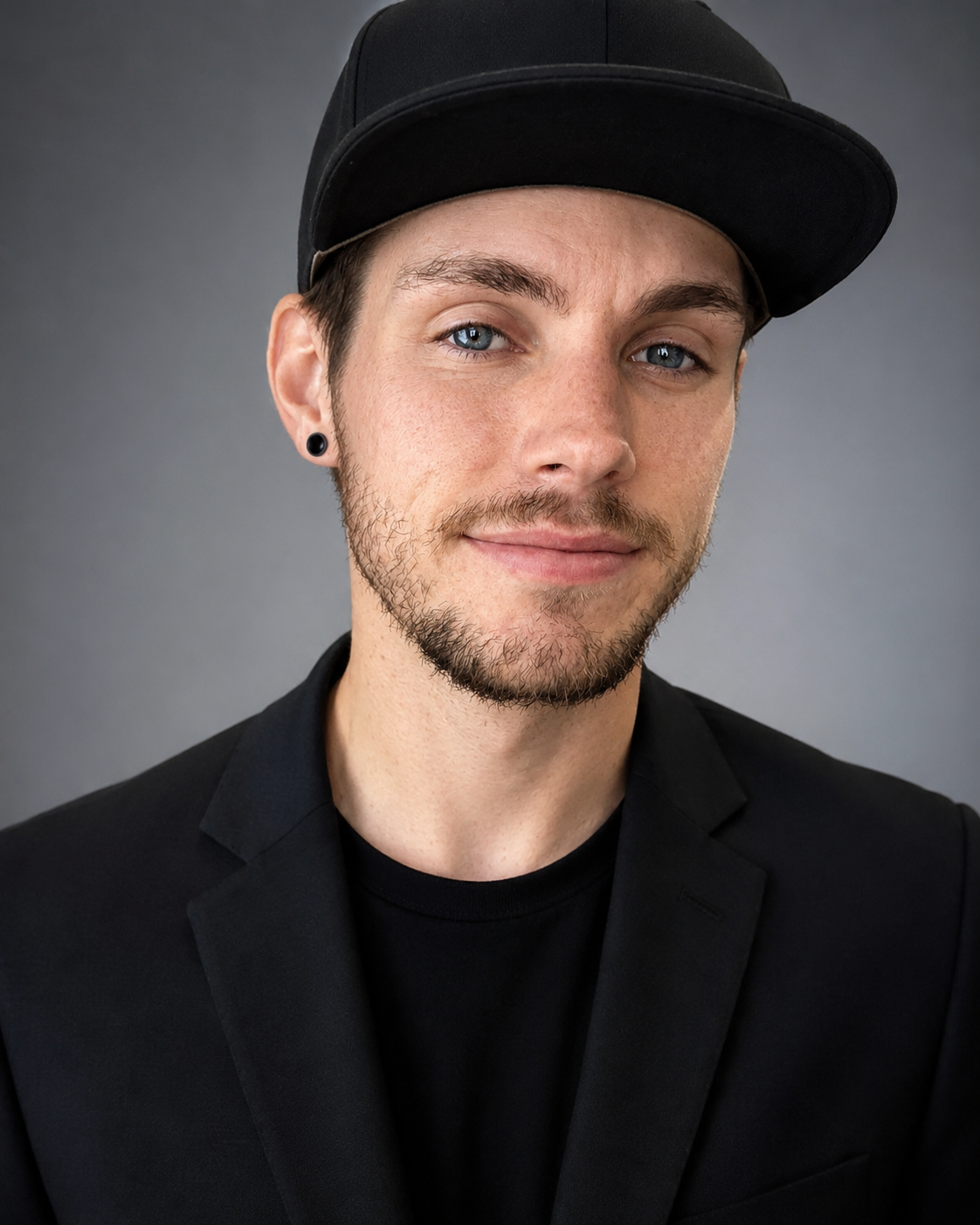 Studio portrait of a person in a black cap and blazer, with a slight smile against a gray background