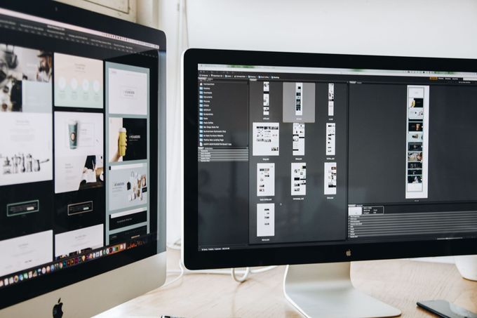 Two desktop monitors showing design layouts on a desk in a bright workspace
