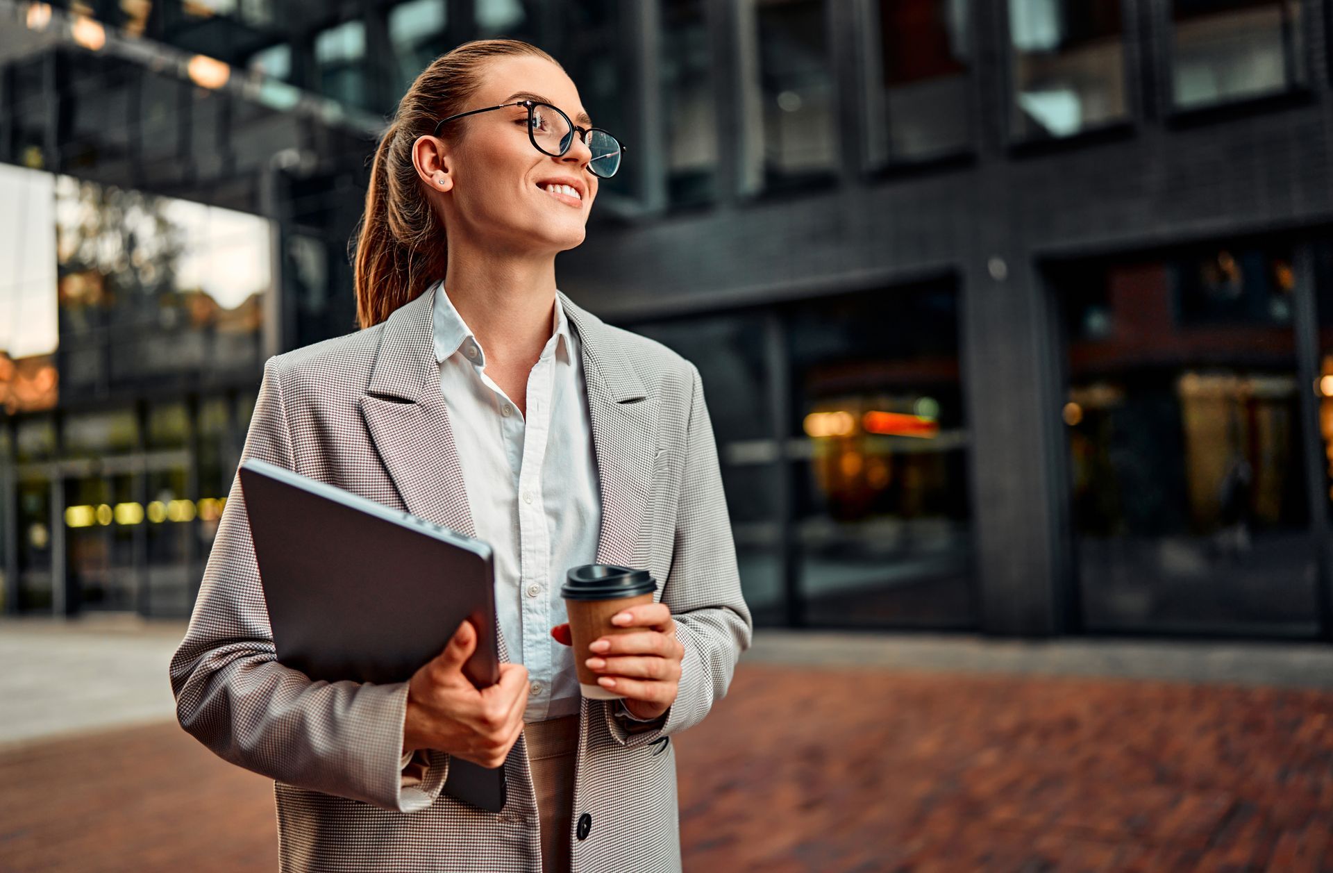 Woman in blazer holding laptop and coffee, smiling outside a building.
