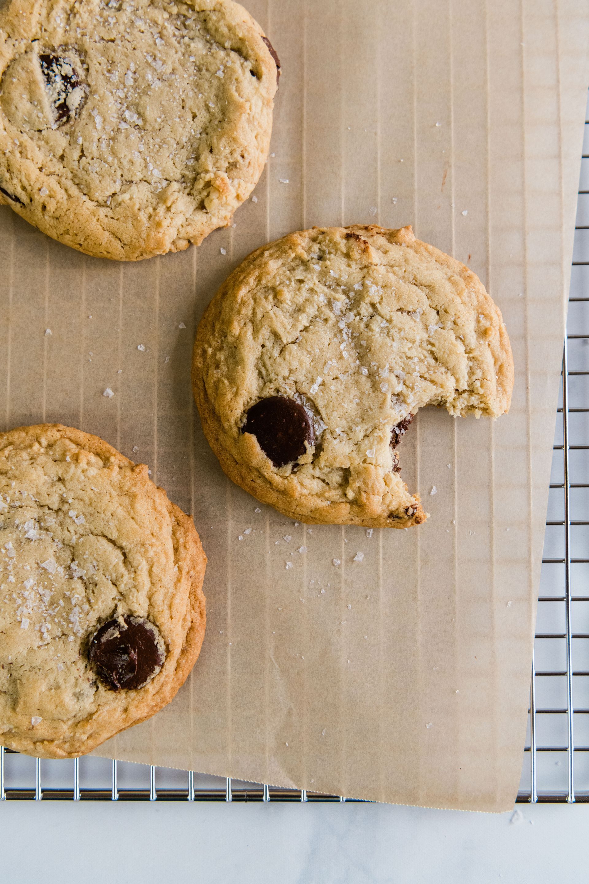 Three chocolate chip cookies are sitting on a cooling rack with a bite taken out of one of them.