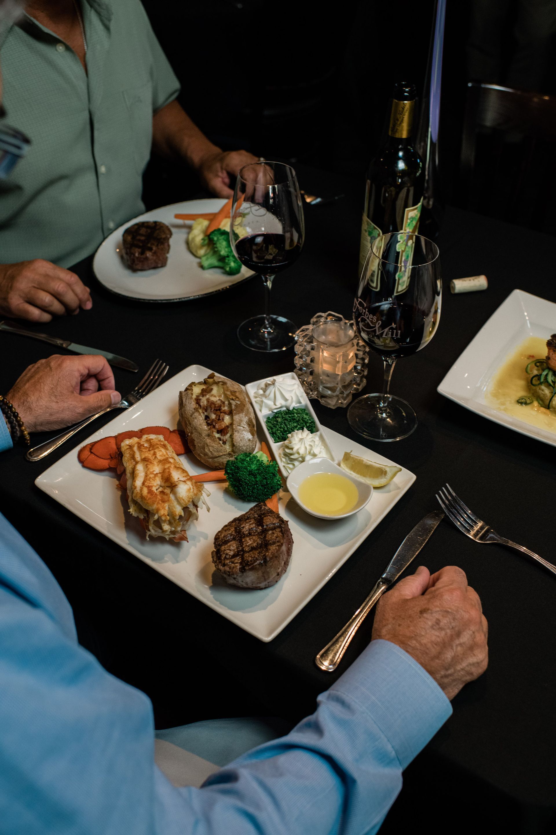Two men are sitting at a table with plates of food and wine.