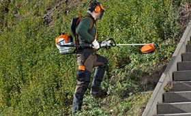Persona che utilizza un decespugliatore a zaino su un pendio erboso accanto alle scale, indossando dispositivi di sicurezza.