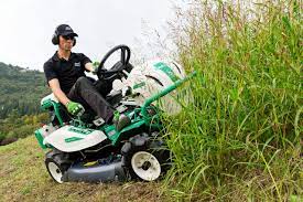 Uomo che utilizza un tosaerba verde da passeggio in collina, tagliando l'erba alta all'aperto.
