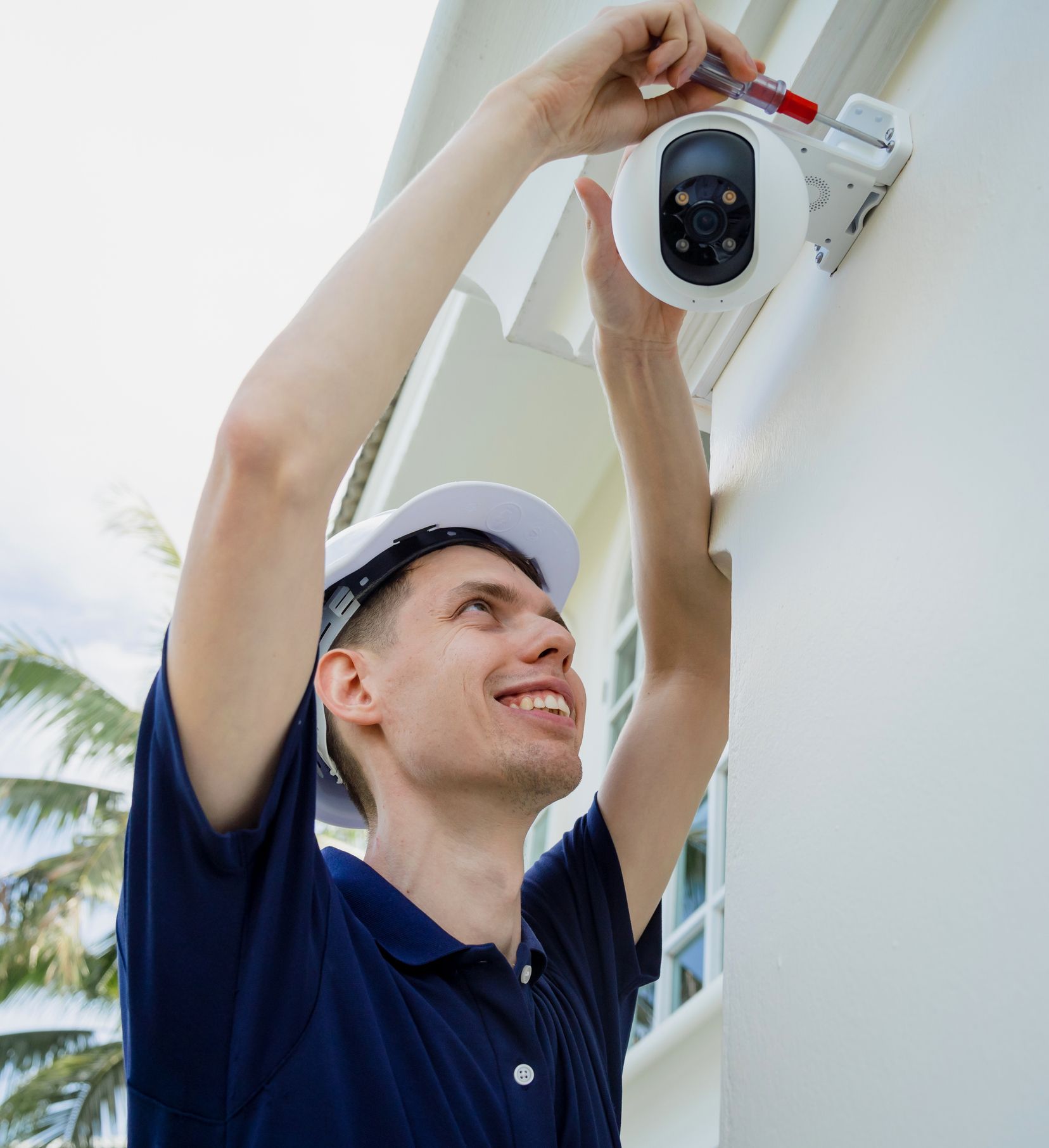 Smiling man in blue shirt installs a security camera on a white building exterior.
