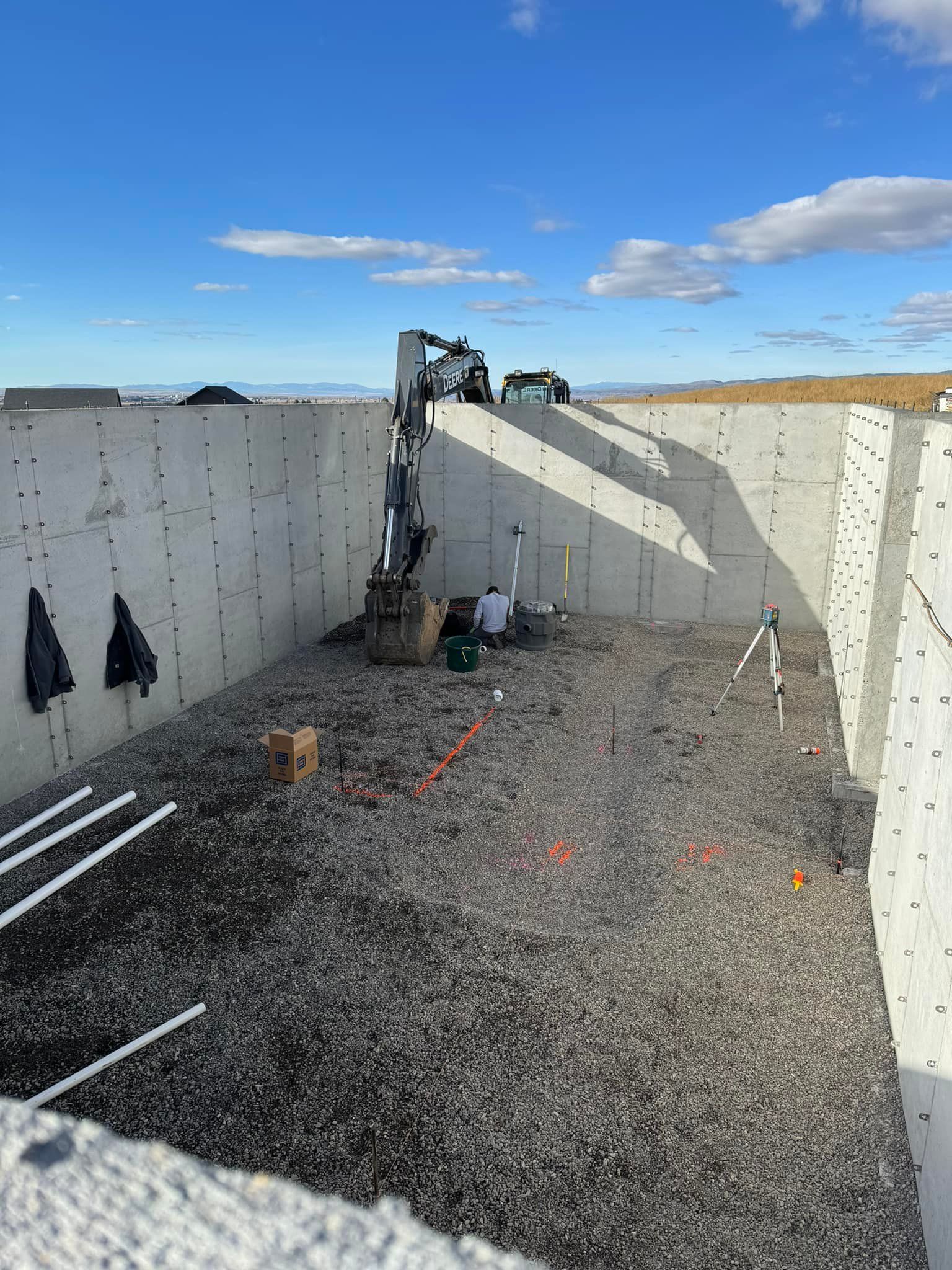Construction site with excavator and workers in a concrete foundation pit filled with gravel, under a blue sky.