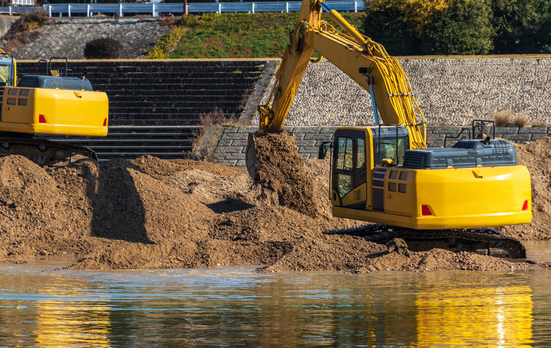 Yellow excavator digging in water, scooping mud; other excavator visible in the background.