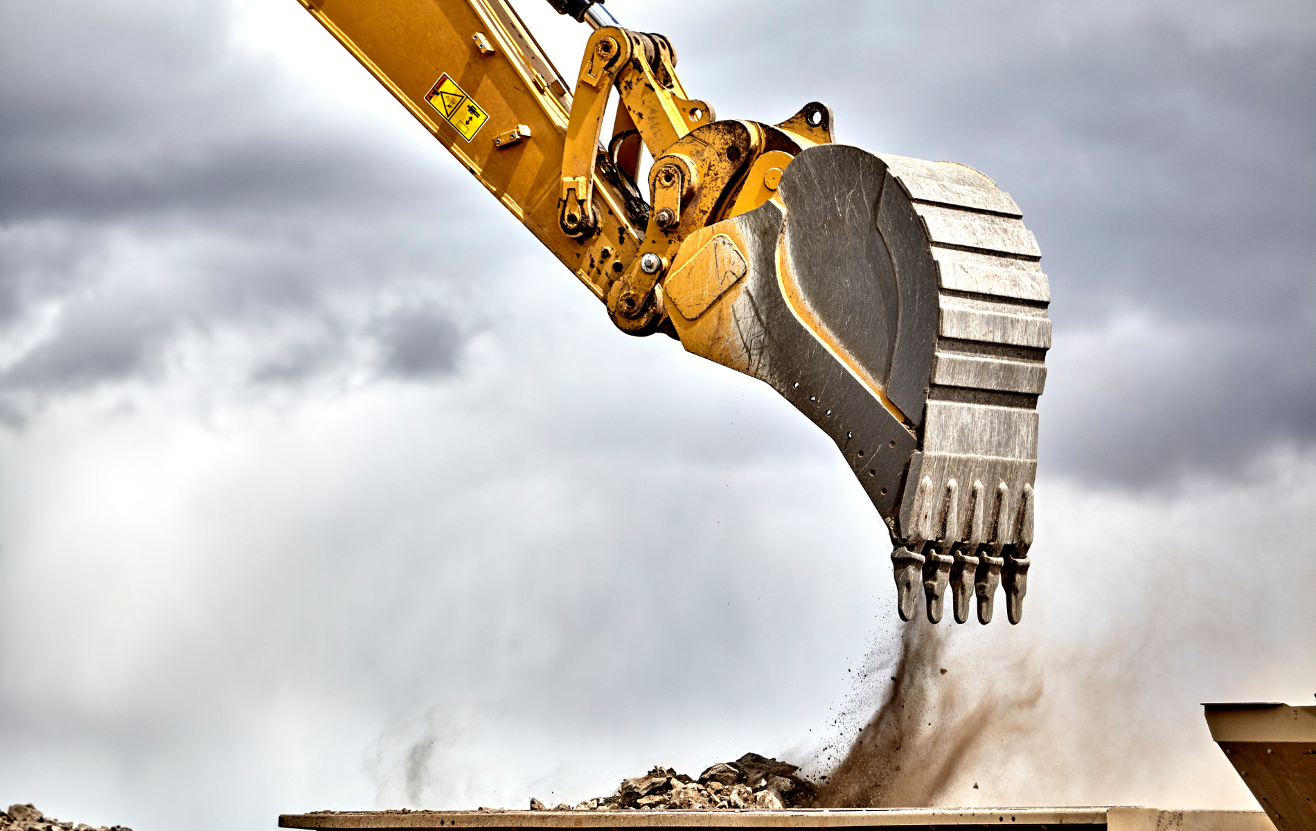 Yellow excavator bucket dumping debris against a cloudy sky.