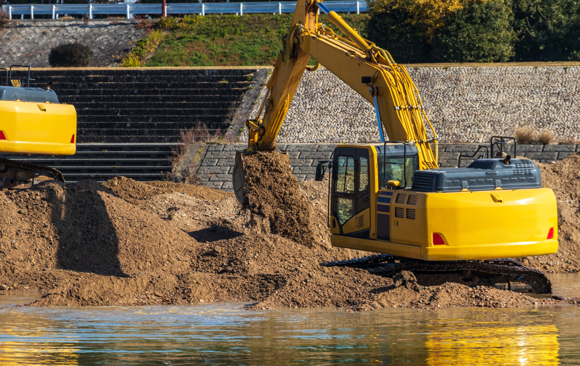 Yellow excavator digging in water near a stone wall.
