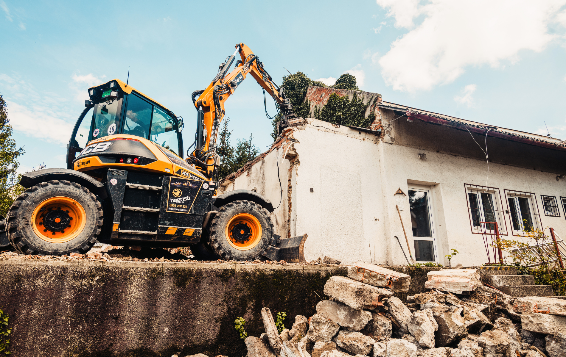 Yellow excavator demolishing a white building; debris pile in foreground, sunny day.