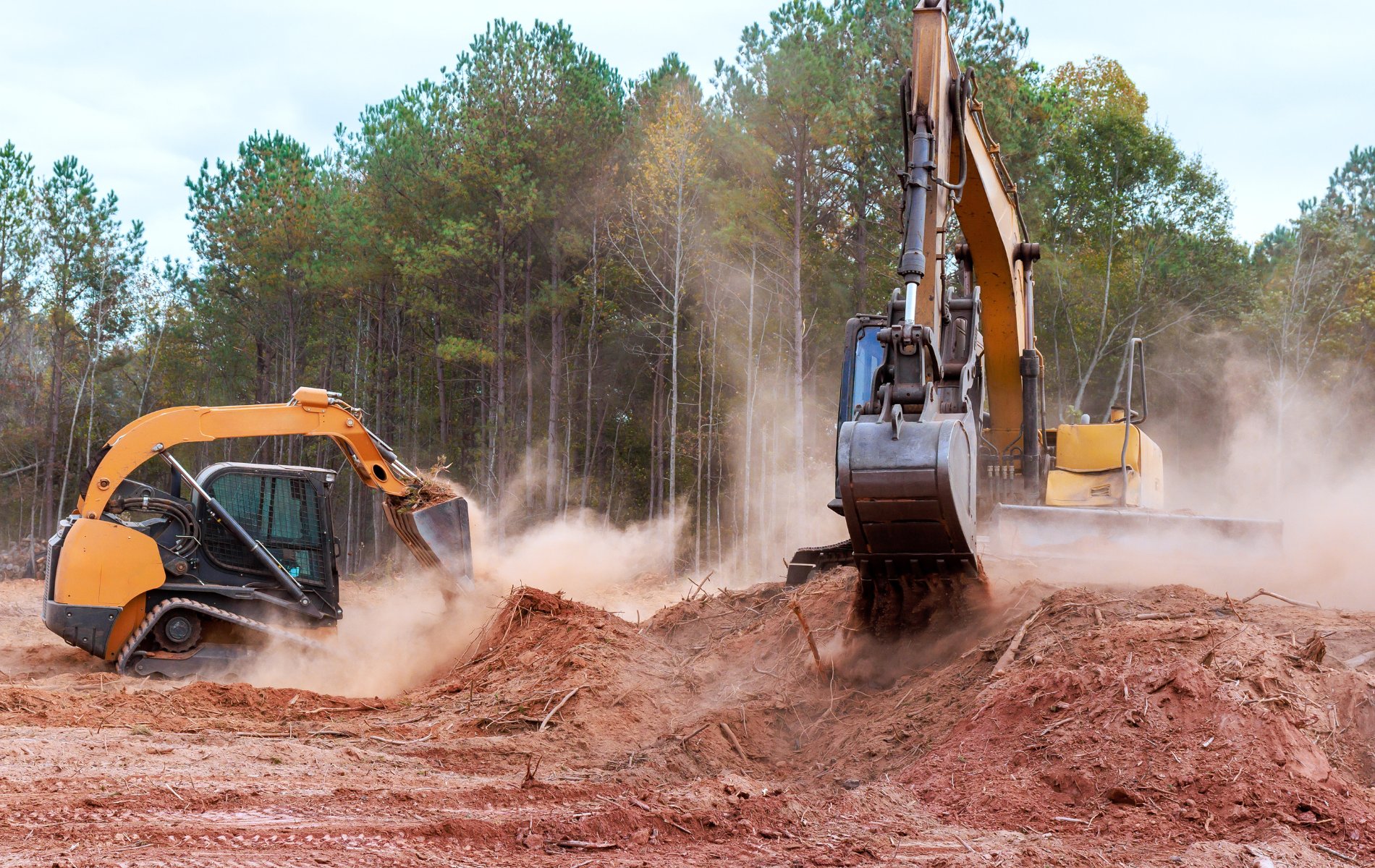 Two yellow excavators digging in dirt, trees in the background, creating dust.
