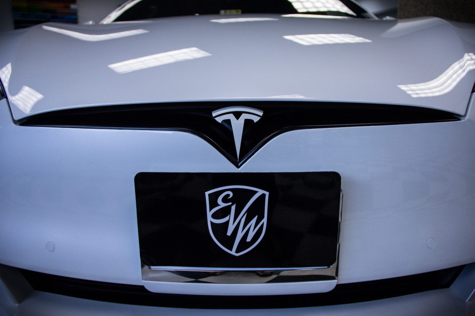 White Tesla car front with license plate; Tesla logo above, EV logo on plate.