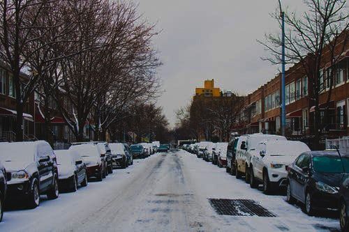 Snow-covered street with parked cars, bare trees, and brick buildings under a cloudy sky.