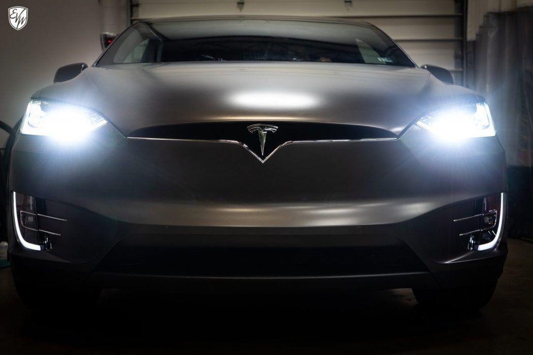 Front view of a Tesla Model X with bright headlights. Gray matte finish in a dimly lit garage.