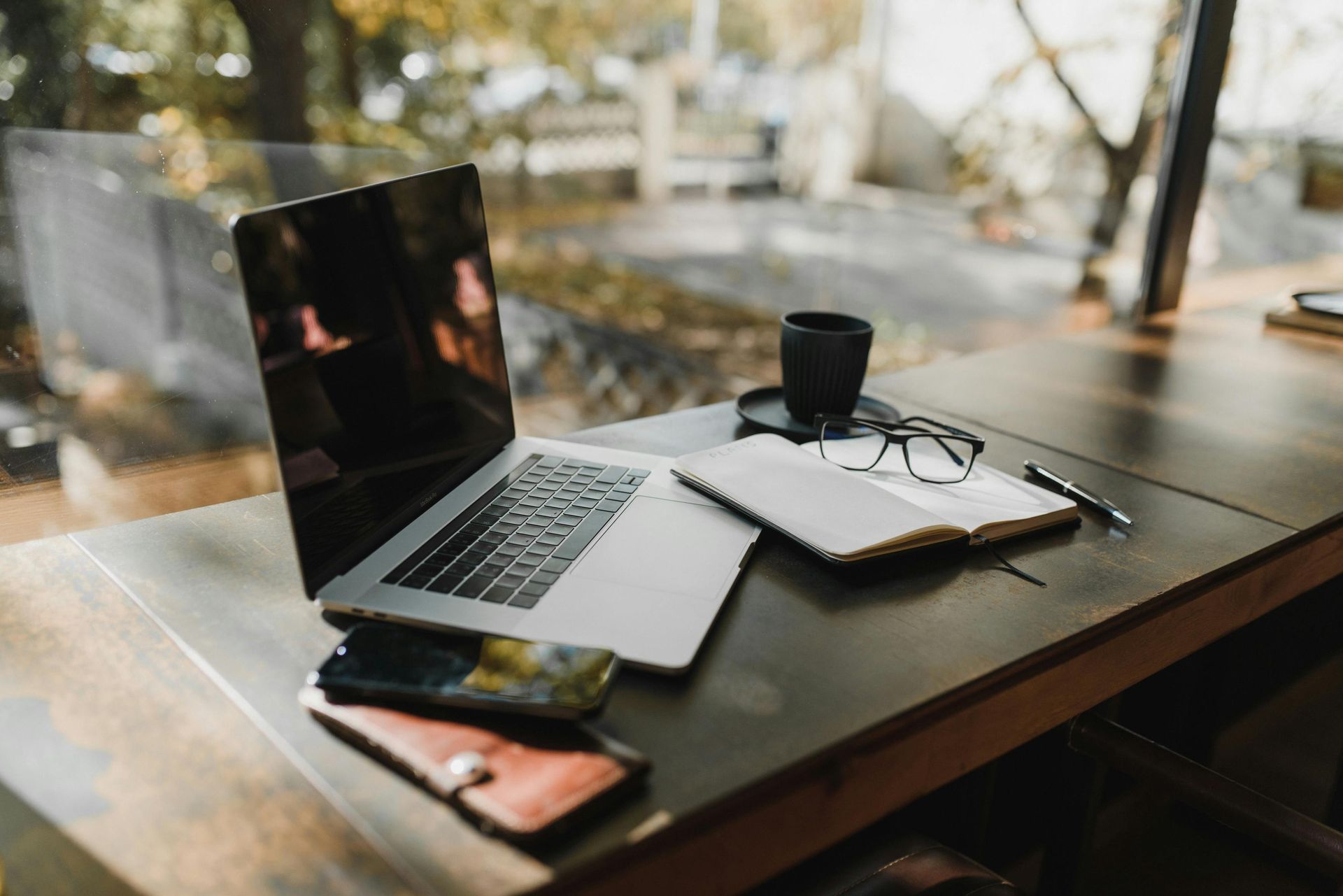 Laptop, notebook, glasses, and coffee cup on a wooden table near a window.