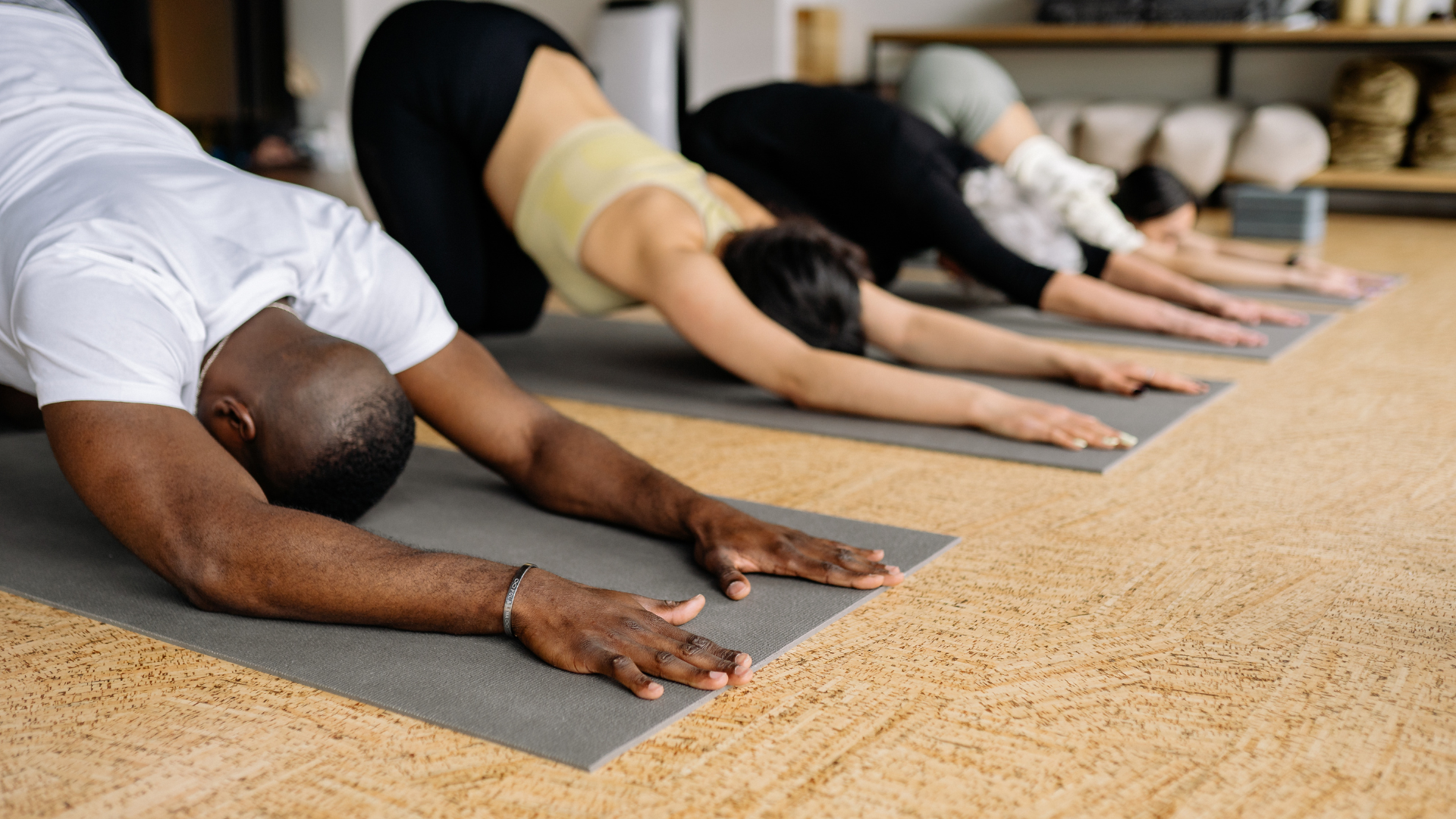 People in yoga class doing child's pose on mats, hands extended, floor is wooden, focus on man in white shirt.