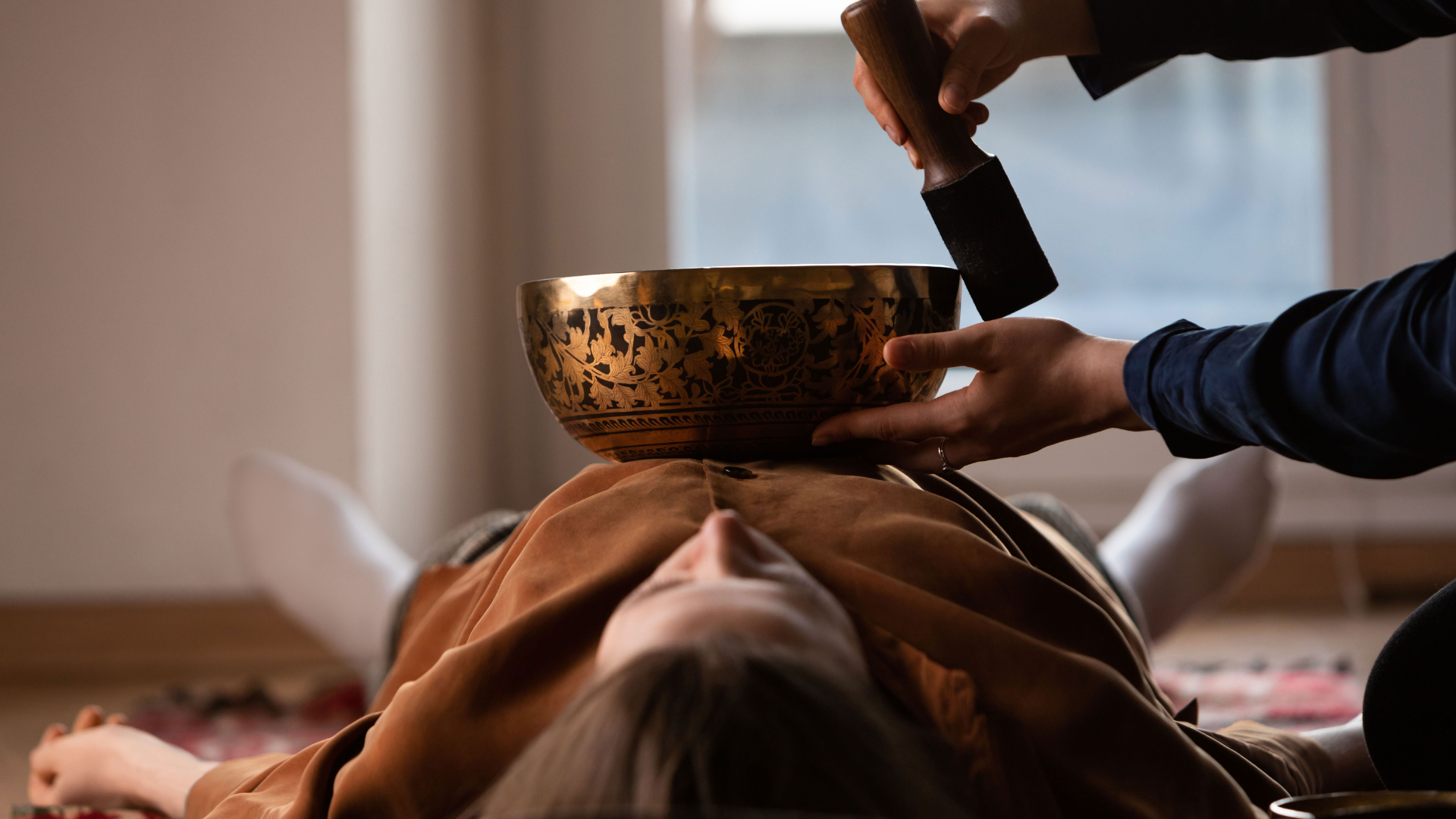 Person receiving a sound bowl therapy session. Bronze bowl over chest, being struck by mallet, indoors.