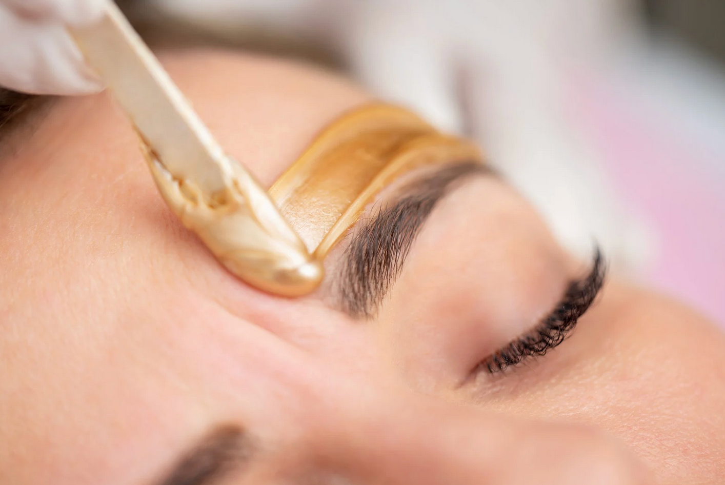 Eyebrow waxing: A person's closed eye while a gloved hand applies wax.
