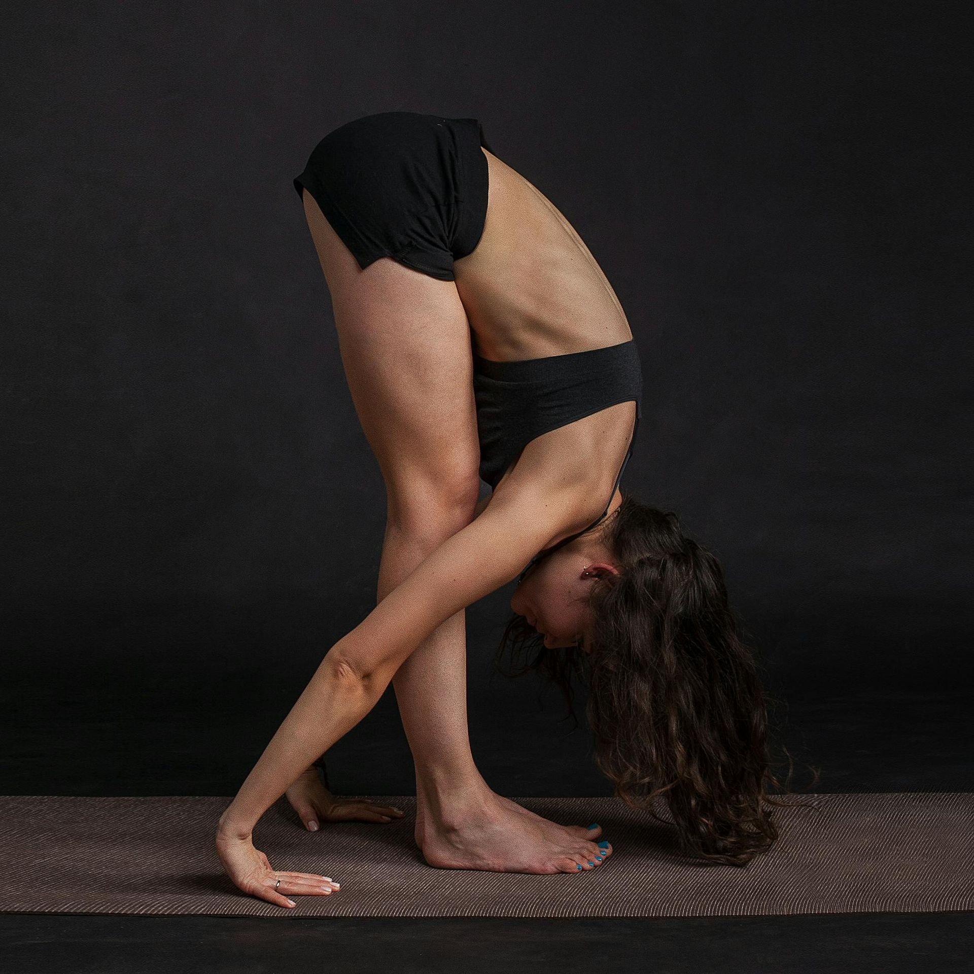 Woman in yoga pose, bending forward with hands reaching toward the floor, dark background.
