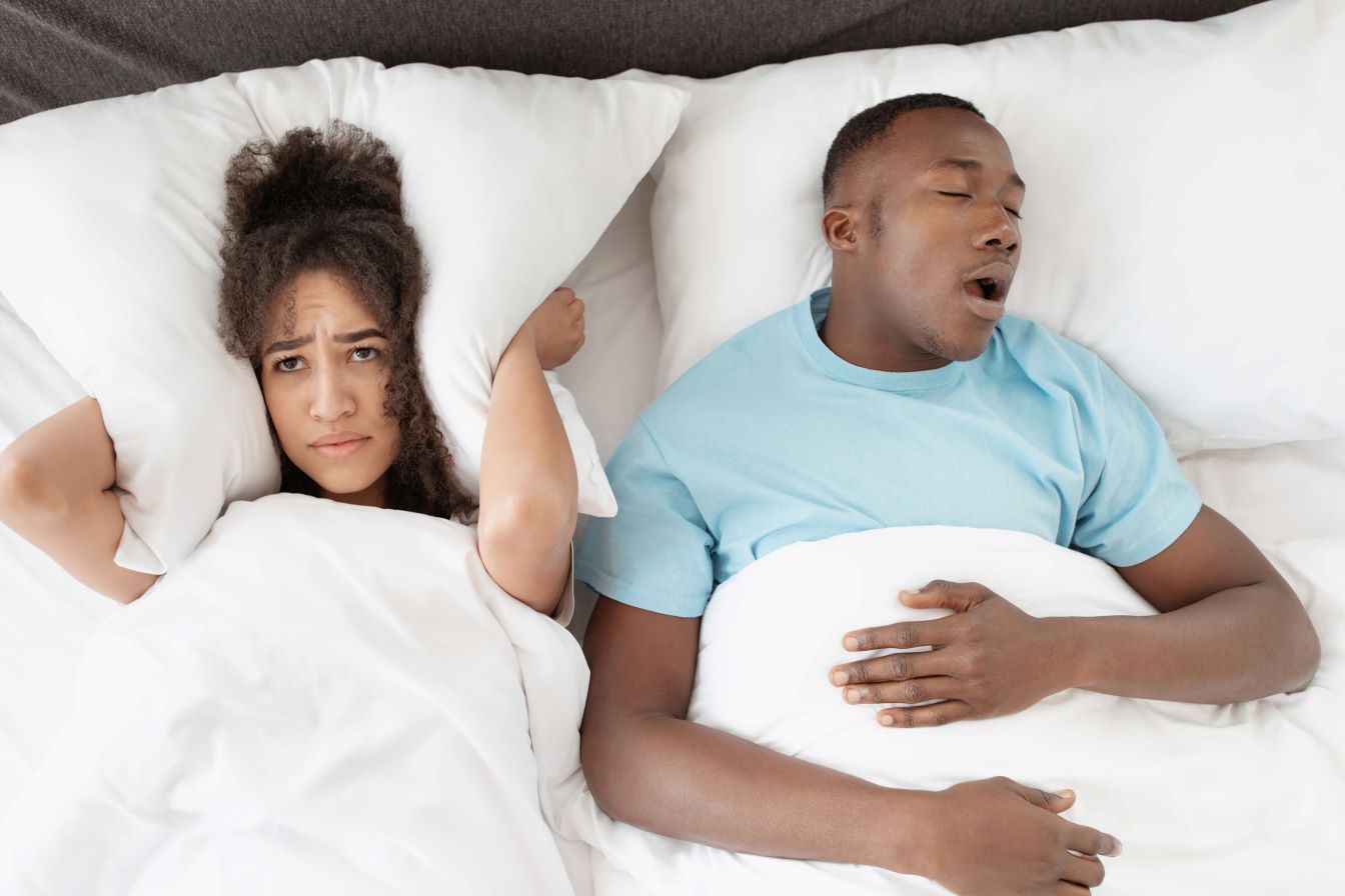 A man is snoring while a woman covers her ears in bed.