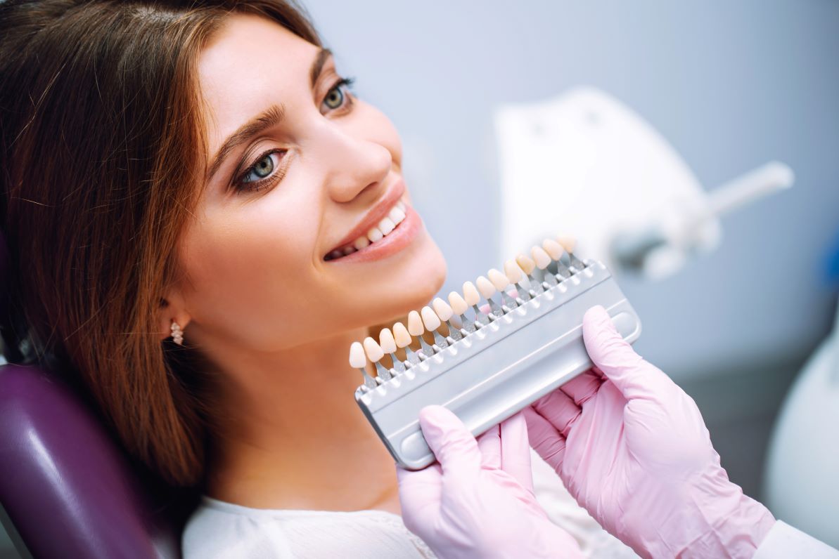 A woman is sitting in a dental chair while a dentist holds a tooth color chart.