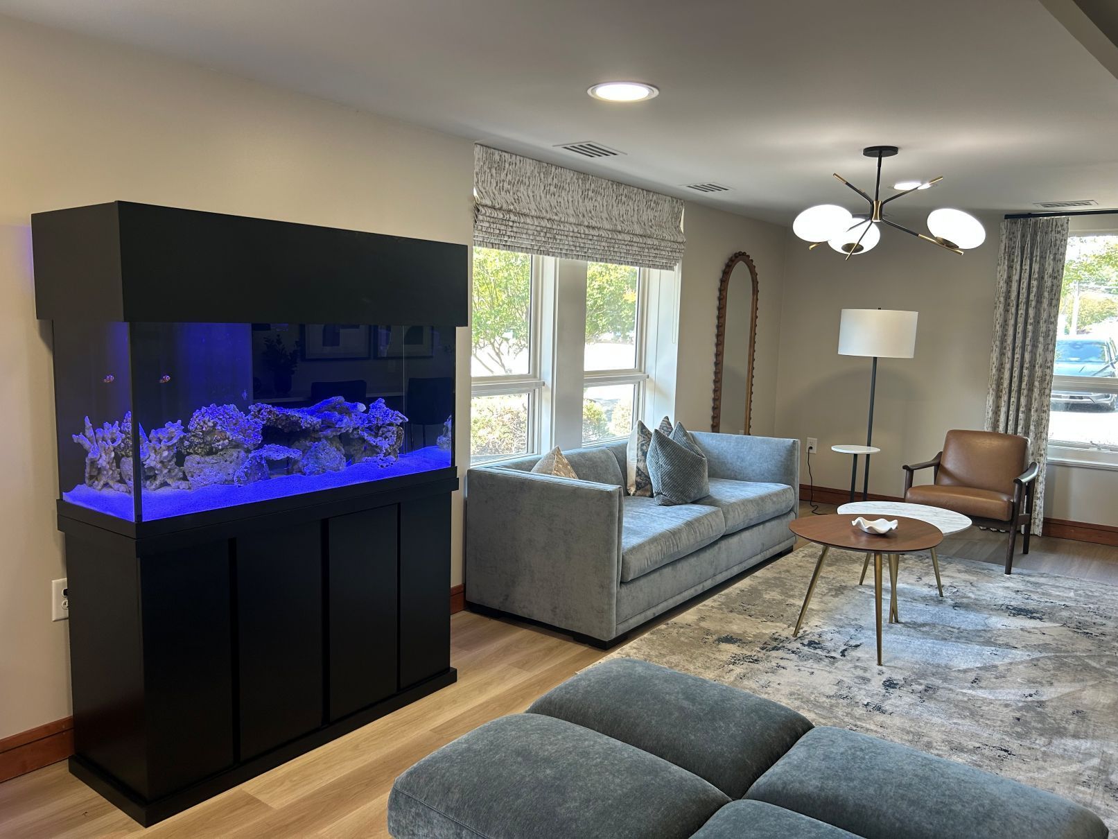 Modern living room with glowing blue aquarium, gray sofa, chair, lamp, and large windows in Raleigh NC