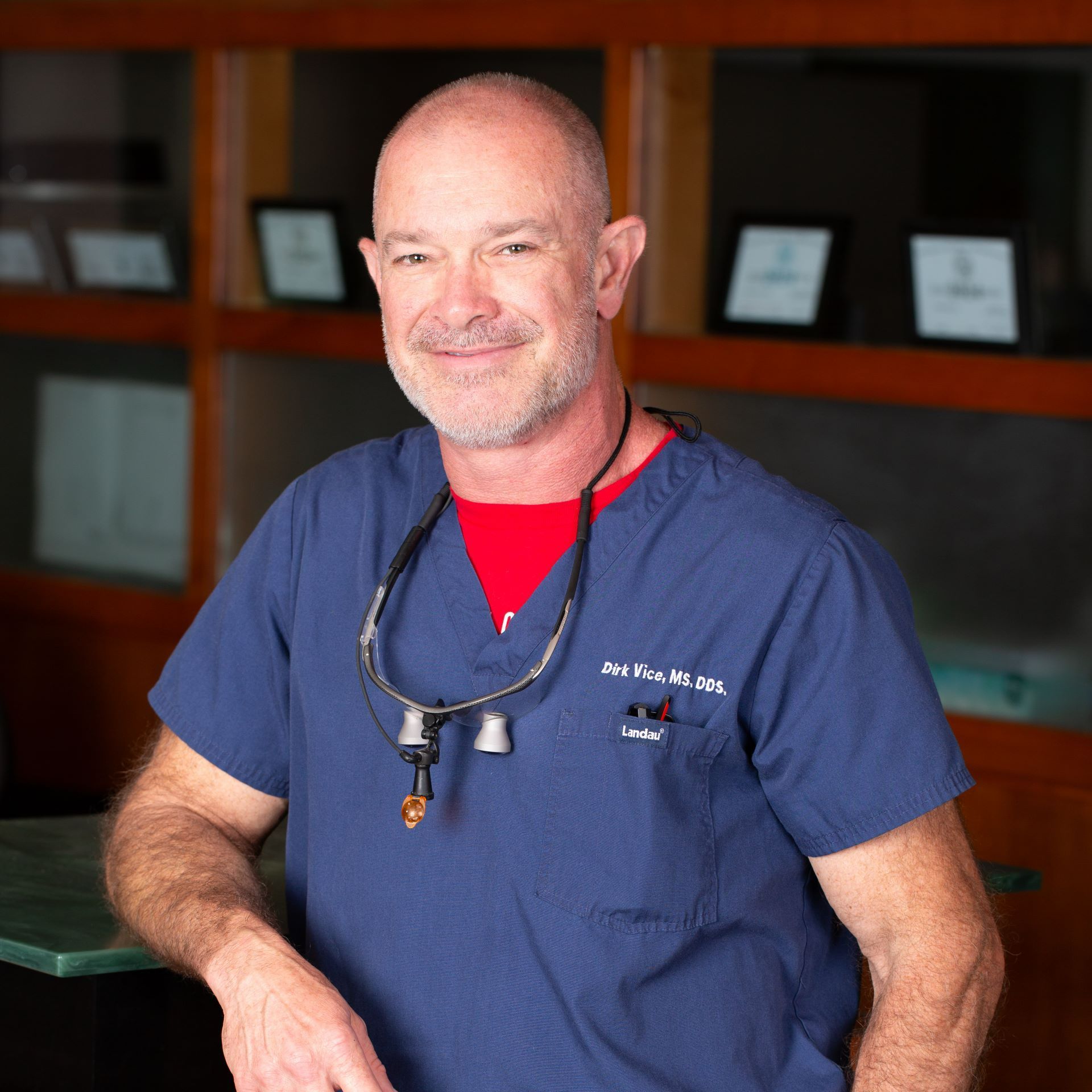 A man wearing a blue scrub top and a red shirt is smiling for the camera.