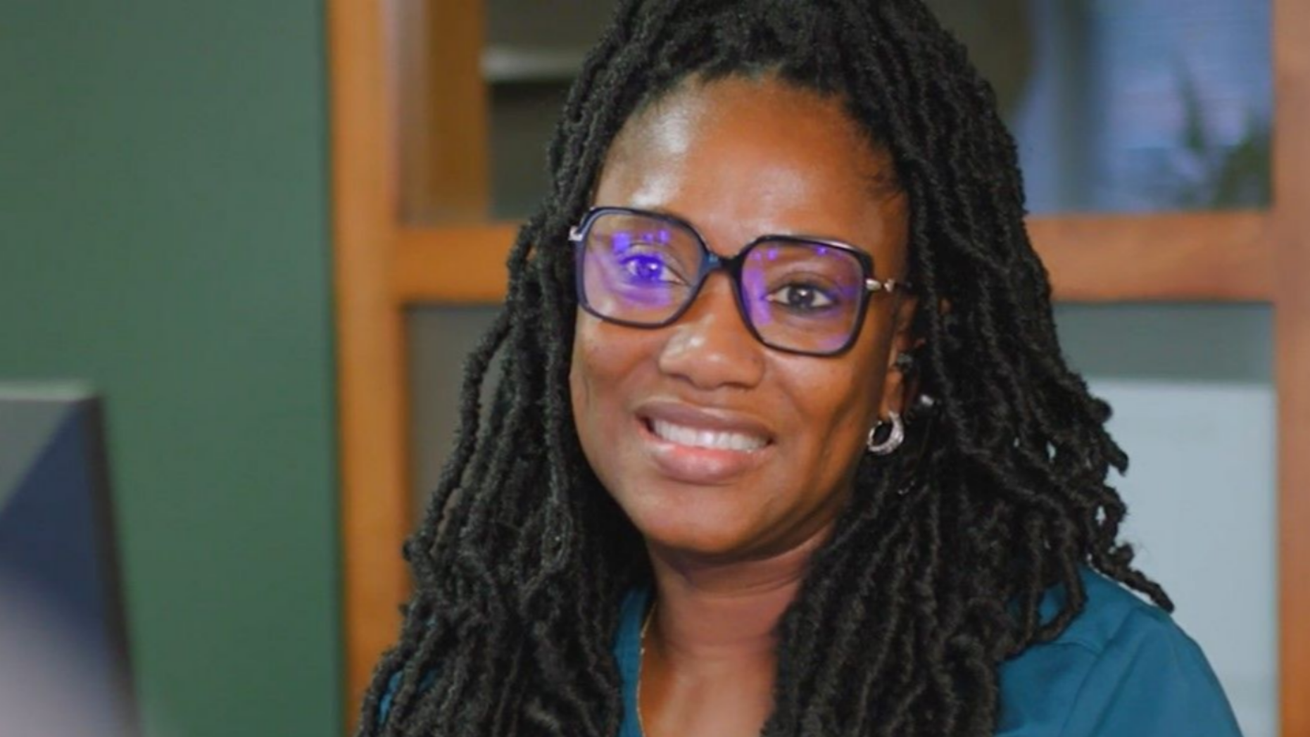 Smiling person with glasses and long braids in a teal top, seated indoors against a softly blurred background in Raleigh, NC.