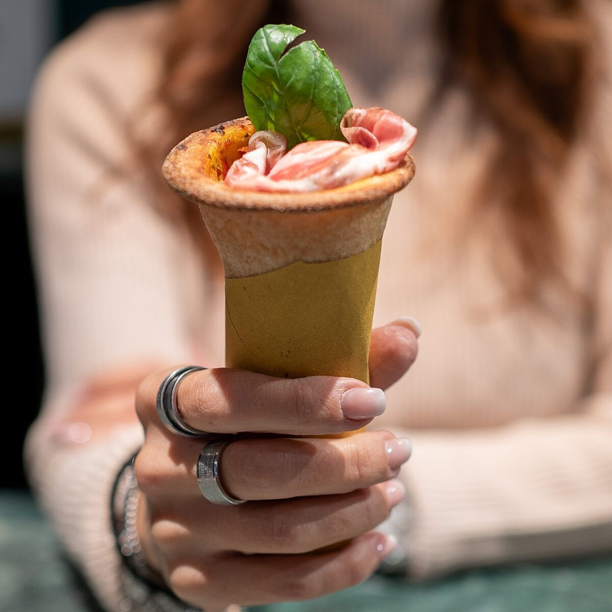 Una mano che regge un dessert a forma di cono guarnito con crema rosa e una foglia di menta, in un'ambientazione sfocata da caffè.