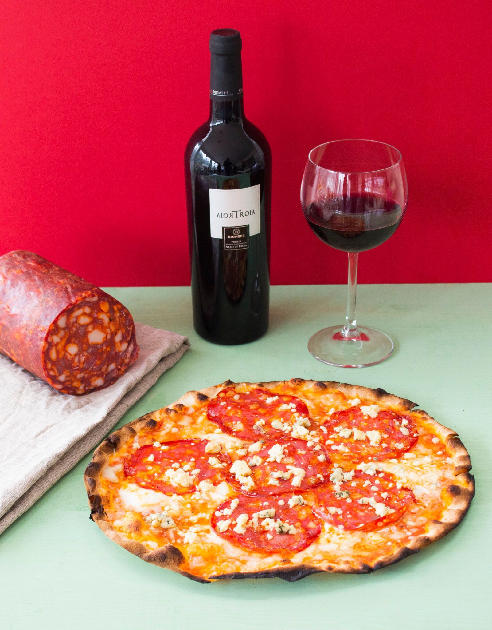 Bottiglia di vino e bicchiere accanto a una pizza e salame affettato su un tavolo con uno sfondo rosso