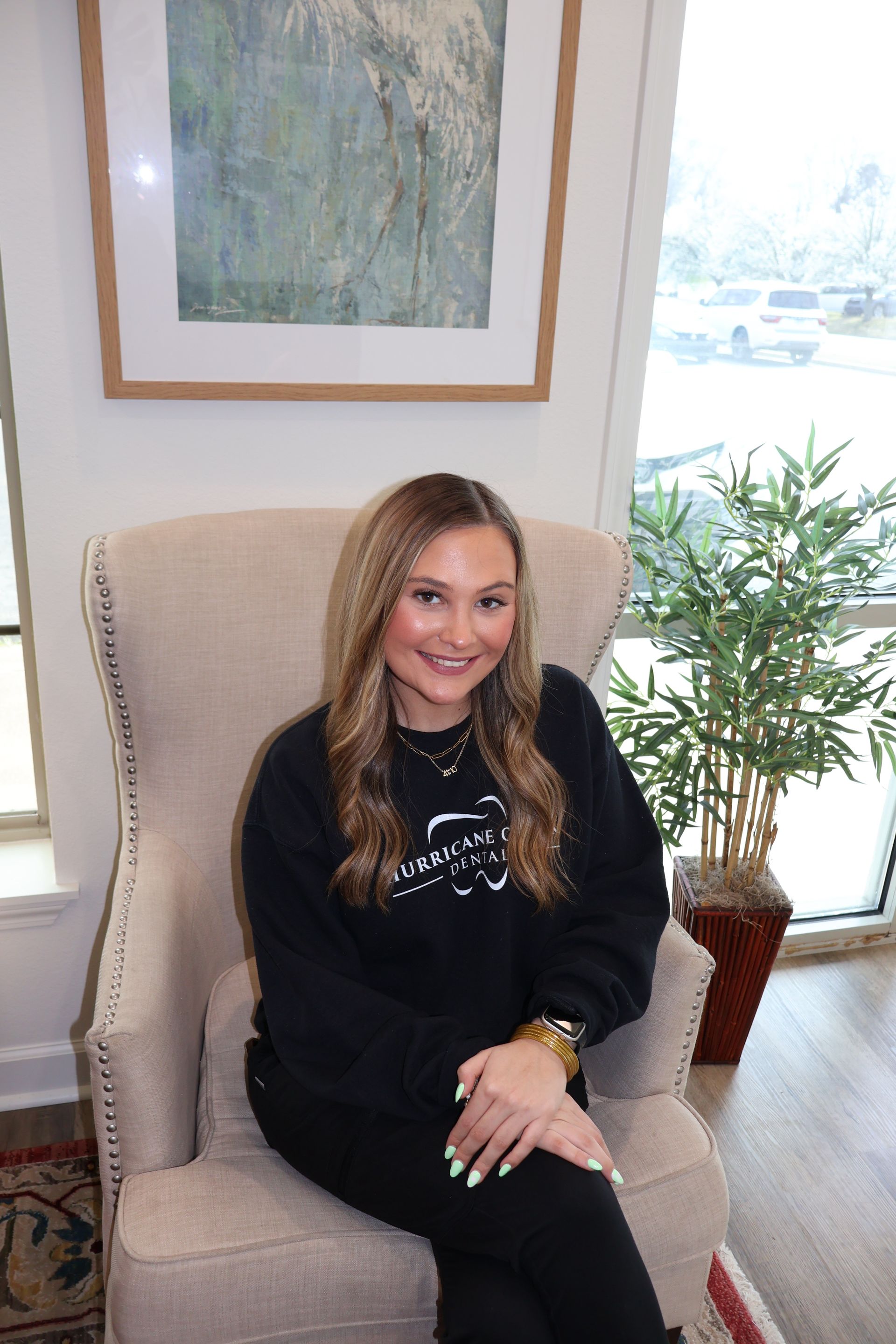 A person sitting in a cream-colored armchair, wearing a black sweatshirt, with a framed painting and plant in the room.