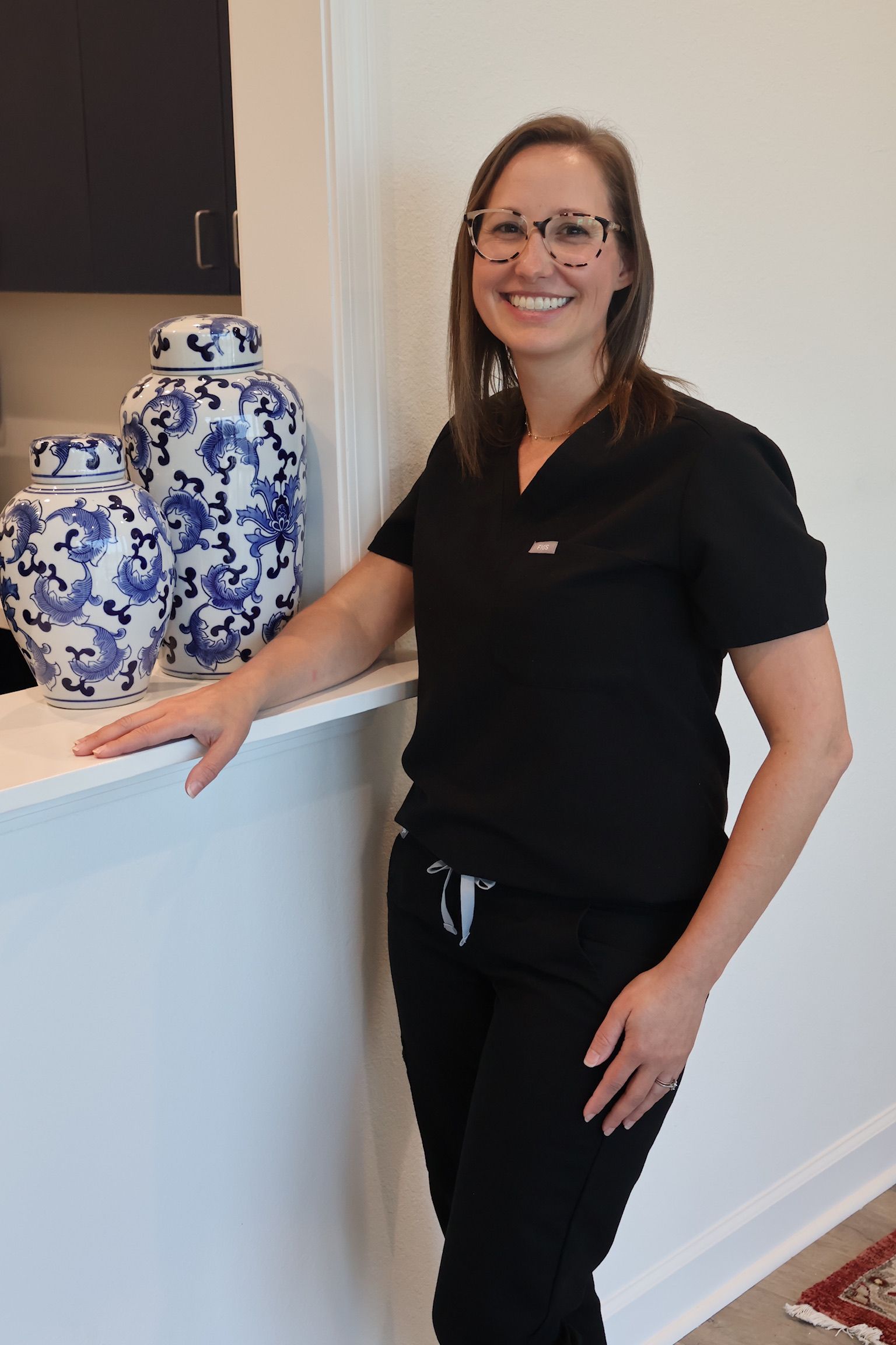 A smiling person in black scrubs stands by a counter with two blue and white decorative ginger jars.