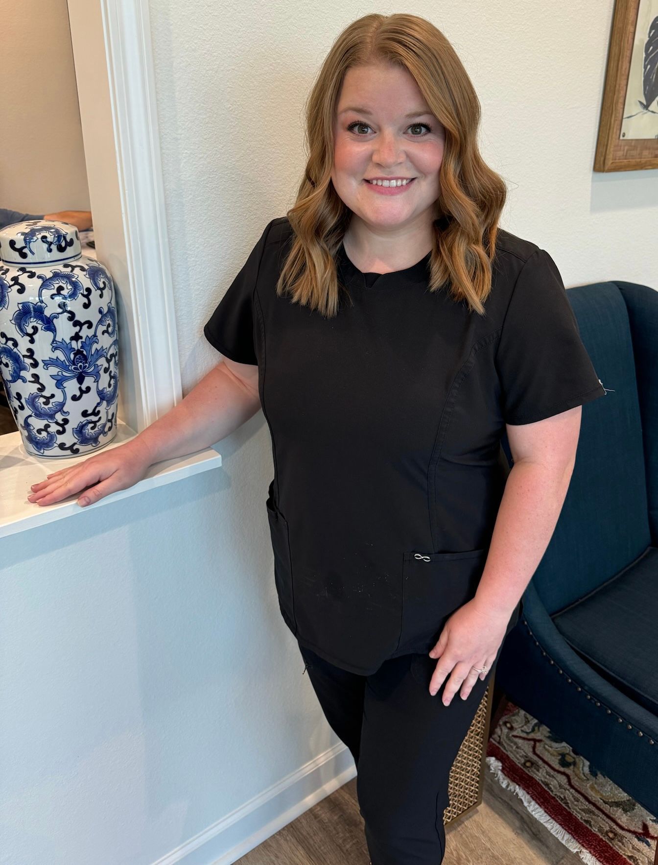 A person with shoulder-length wavy hair, wearing black scrubs, stands smiling in an office setting near a blue vase.