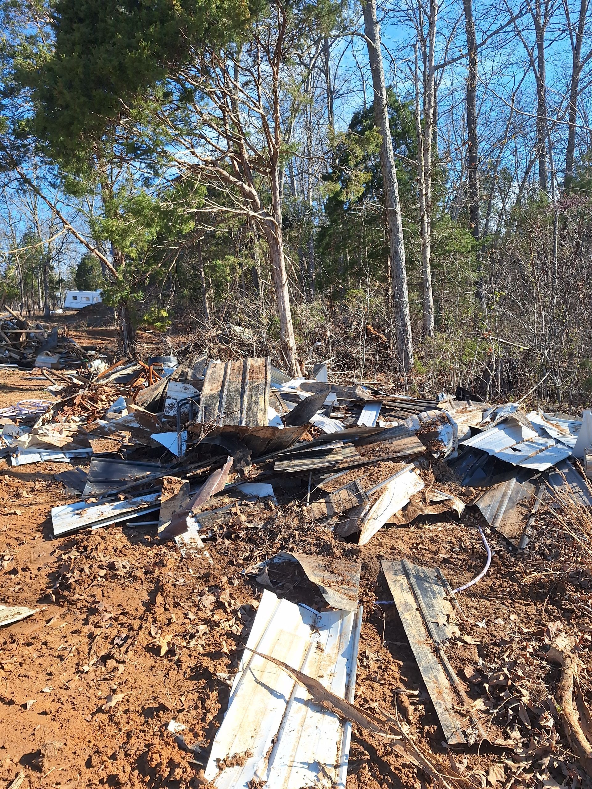 Debris and broken metal scattered on a muddy roadside beside trees under a clear blue sky