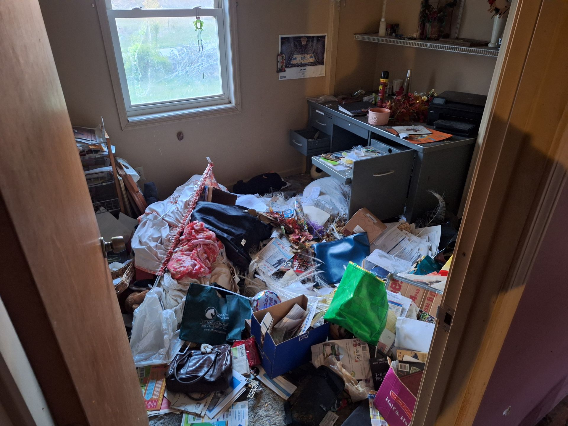 Cluttered bedroom floor covered in piles of trash and clothes, with a desk and window in the background