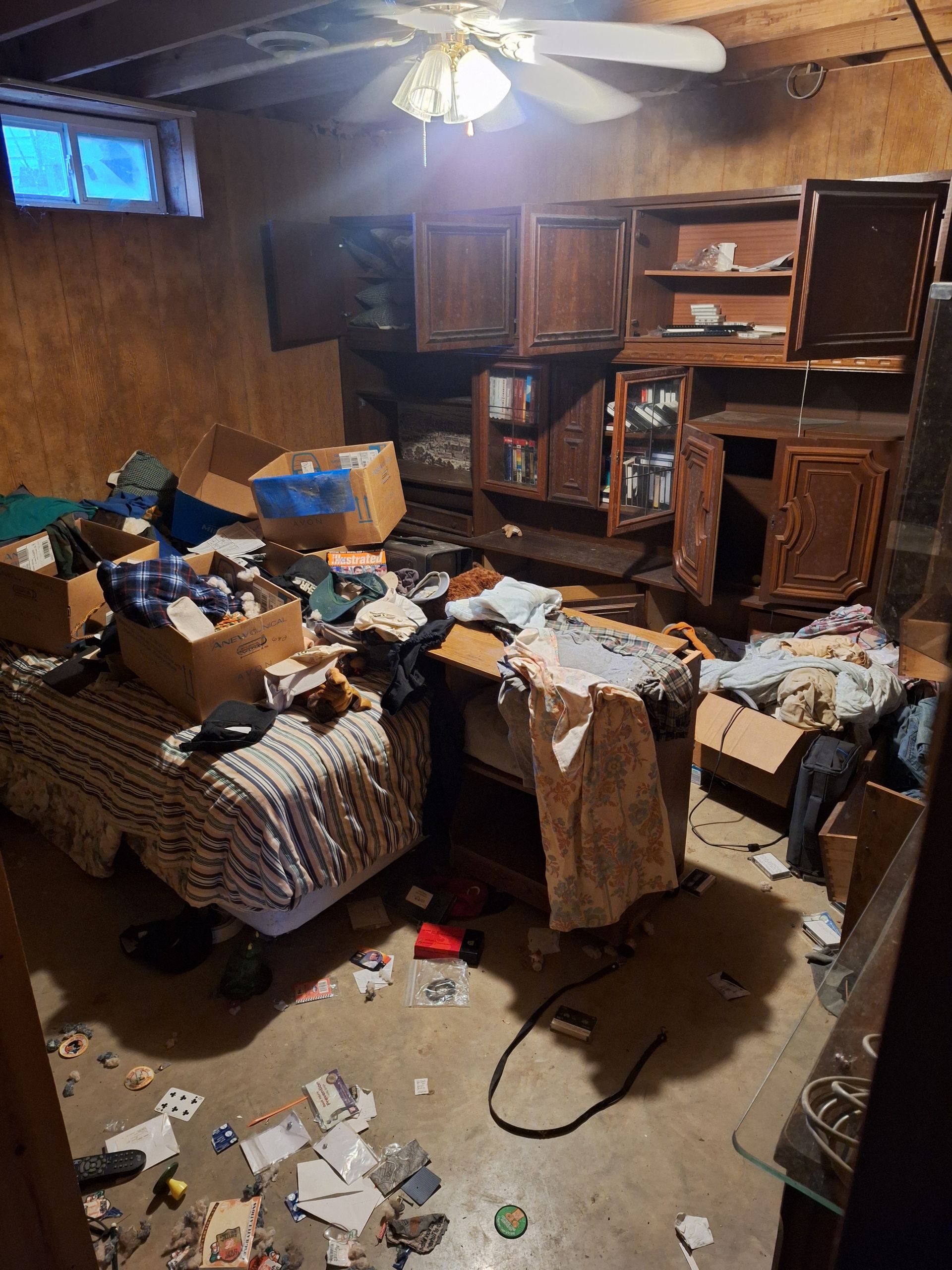 Cluttered, messy room with scattered papers, boxes, and broken furniture under a ceiling fan.