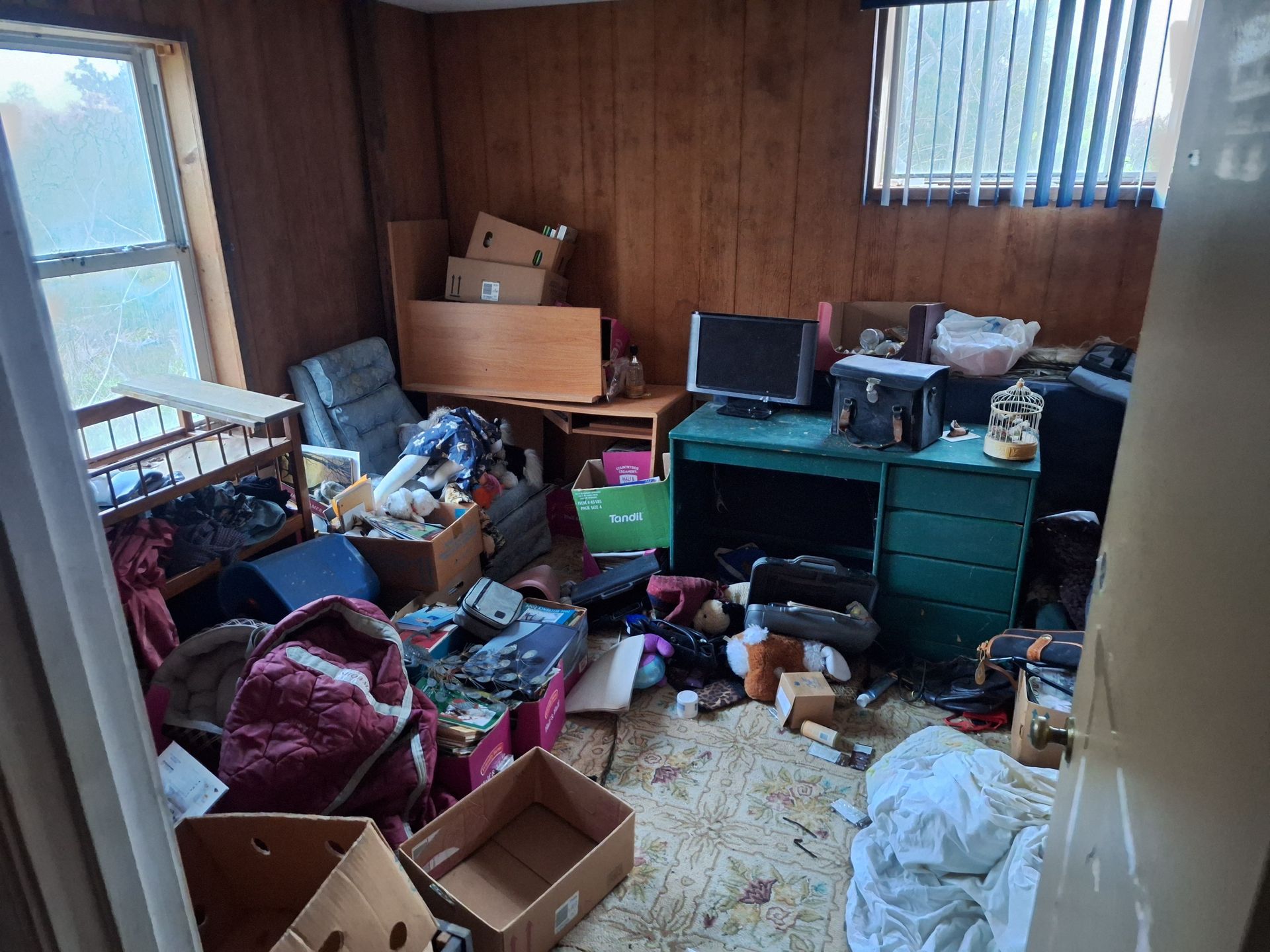 Cluttered small room with boxes, bags, and scattered items beside a desk and window.