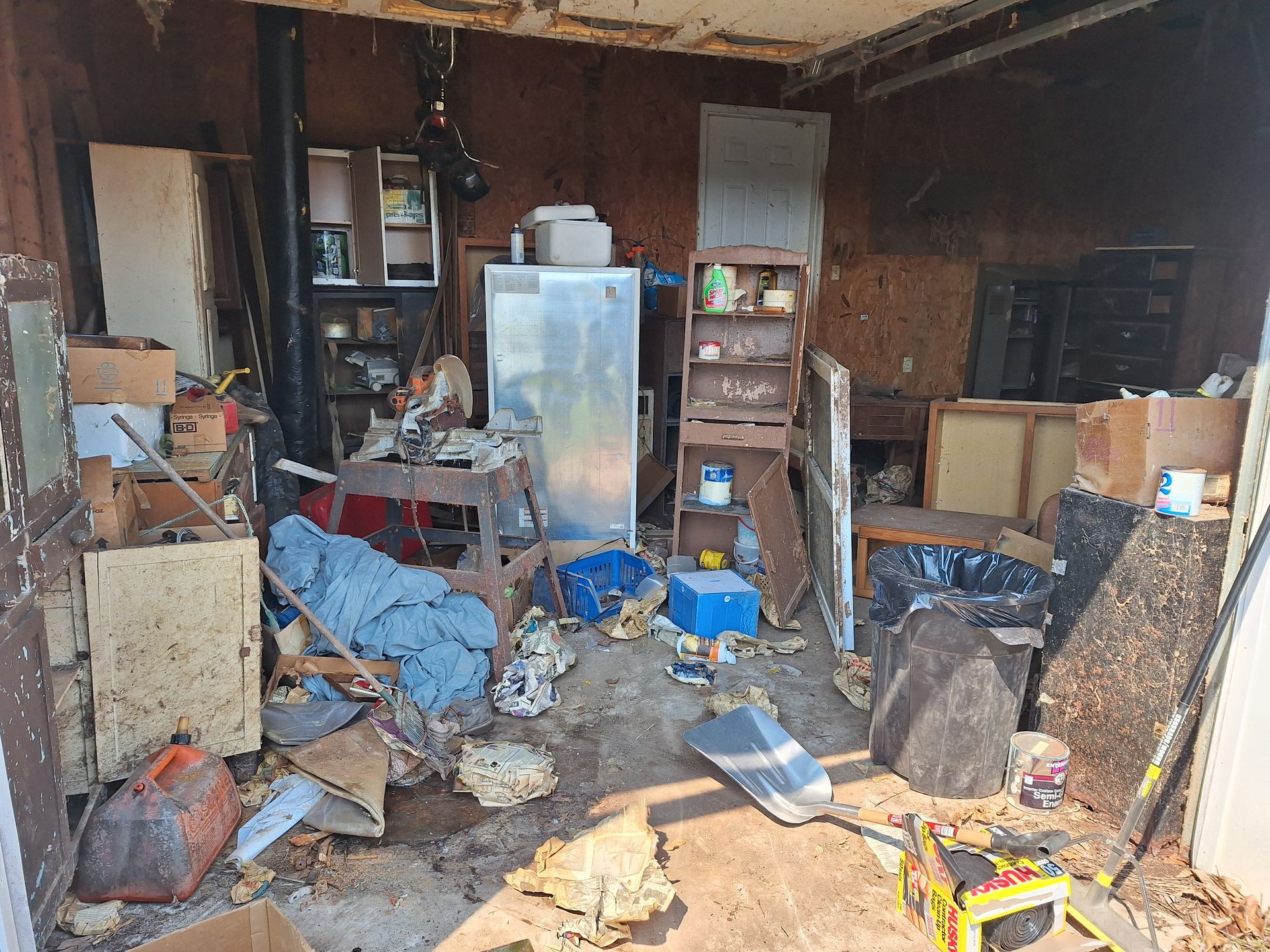 Cluttered, debris-filled room with damaged walls, boxes, bags, and scattered household items.