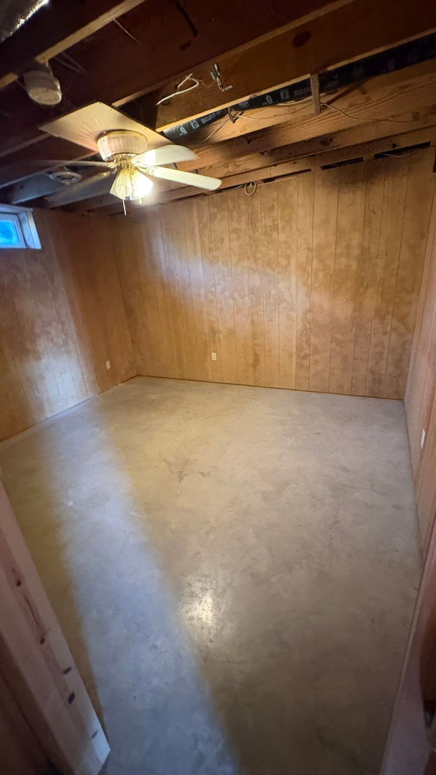 Empty unfinished room with plywood walls, exposed ceiling beams, and a concrete floor under warm light