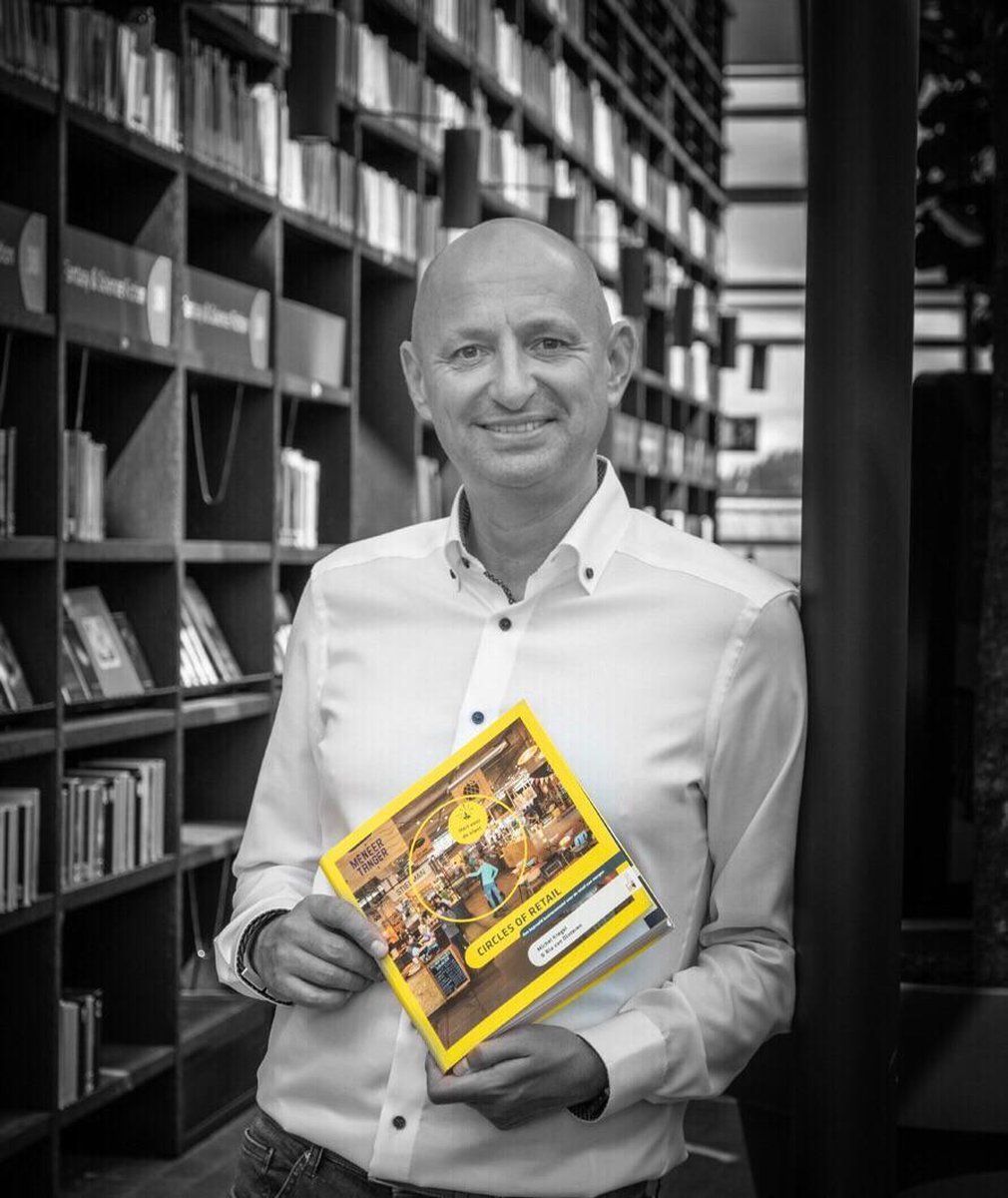 Een glimlachende persoon in een wit shirt staat in een bibliotheek en houdt een boek met een opvallende gele kaft vast.