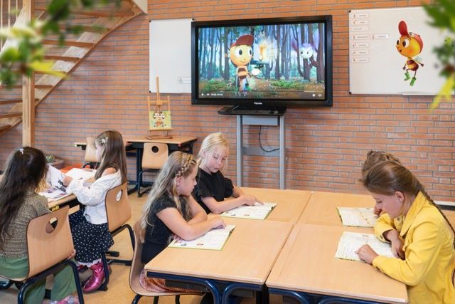 Een groep kinderen zit aan tafels in een klaslokaal en kijkt naar een tekenfilm op een flatscreen-tv.
