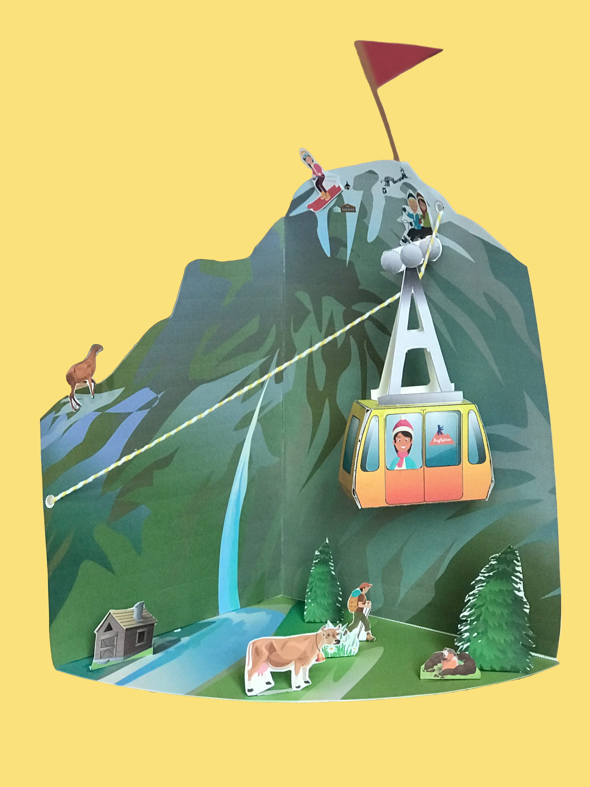 Een papieren berglandschap met een kabelbaan, een koe, een hut, bomen en een vlag op de top.