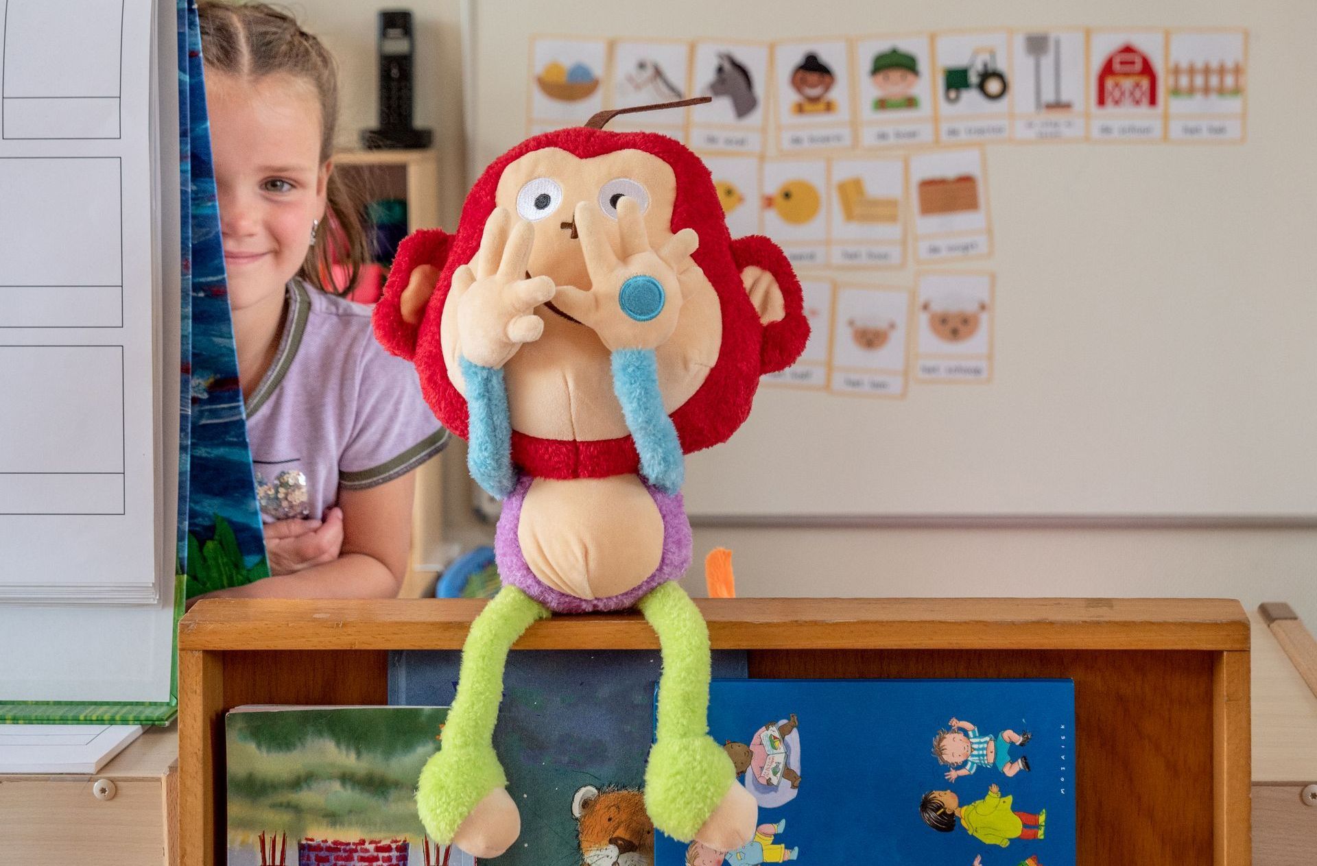 Een klein meisje zit achter een plank met een knuffelaap voor haar gezicht.